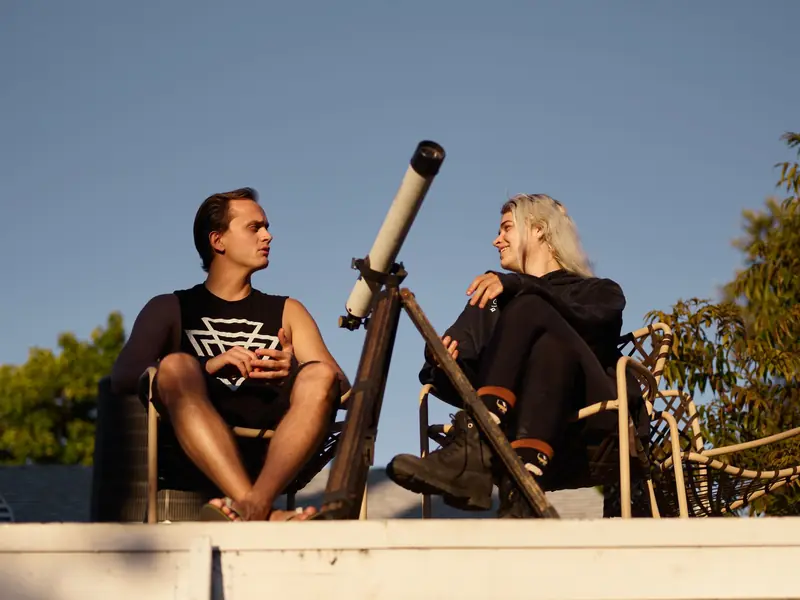 A young couple enjoys a sunny day outdoors, talking and smiling with a telescope nearby