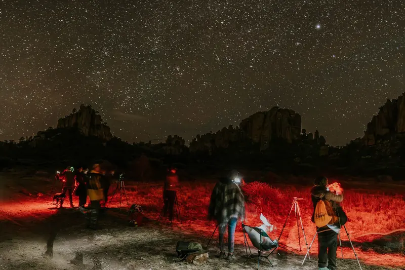 Group of people with cameras taking photos of a starry night sky in a rocky outdoor landscape with red light