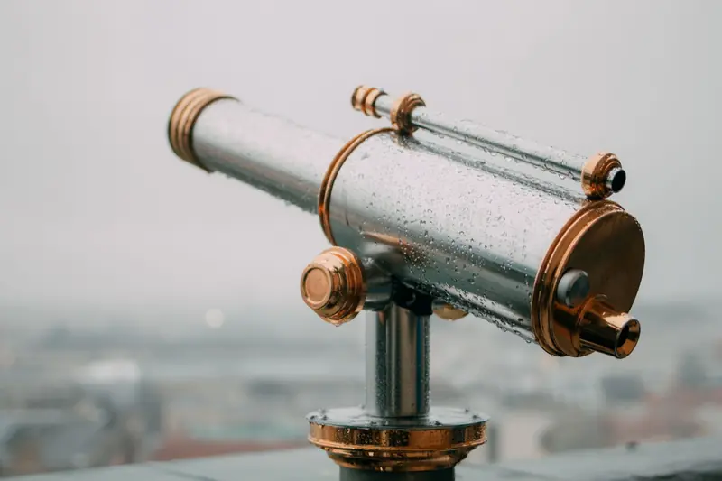 A detailed close-up of a metallic telescope covered in water droplets, set outdoors on a cloudy day